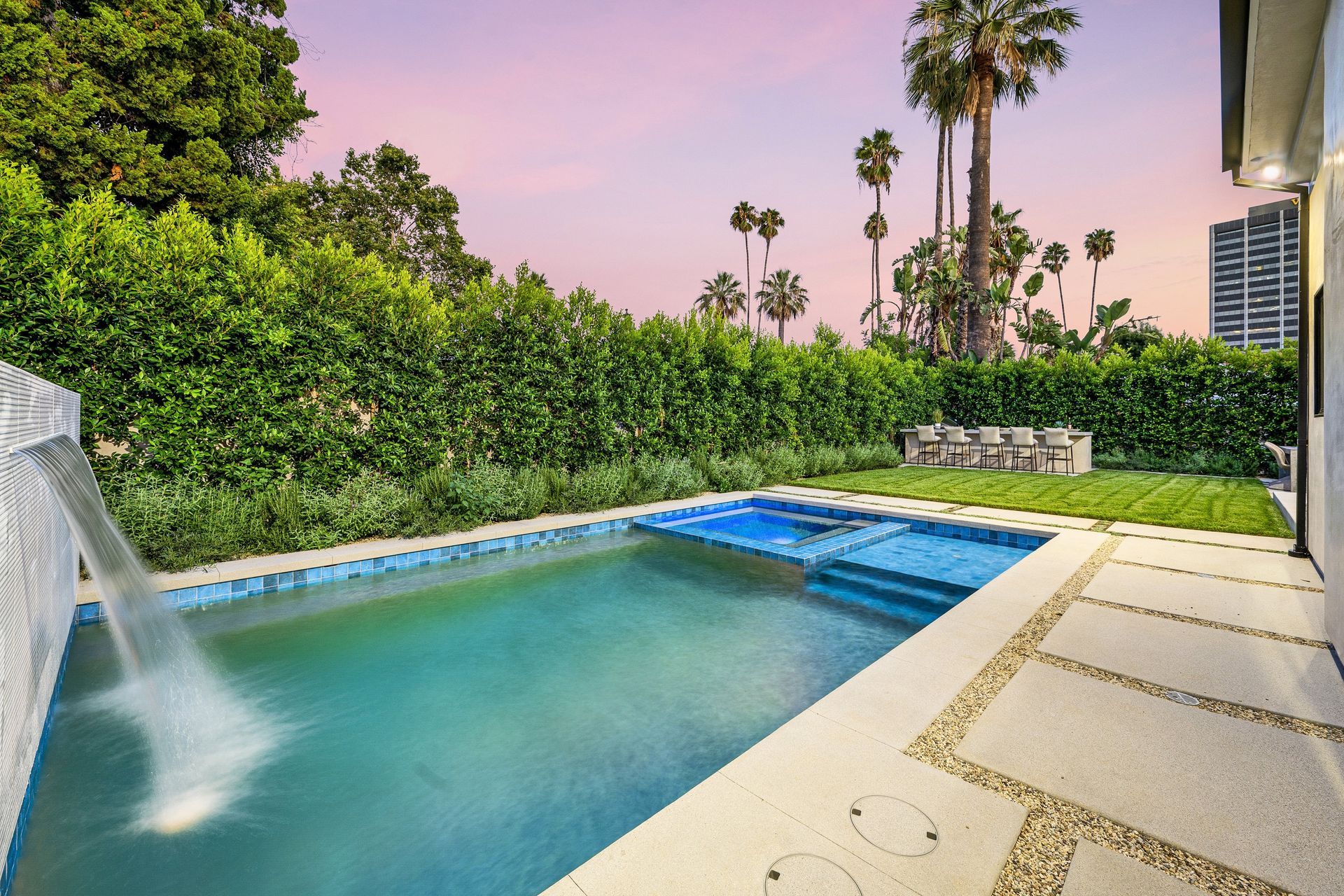 Pool with waterfall, surrounded by greenery, palm trees, and a building. Pink and blue sky.