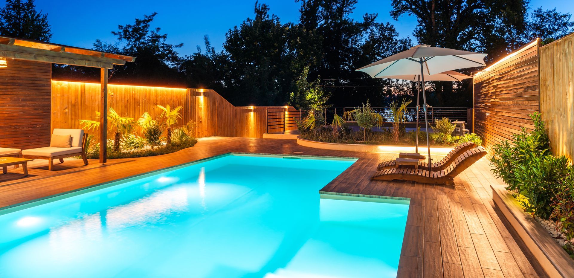 A turquoise pool at night surrounded by wooden decking, trees, and warm lighting.