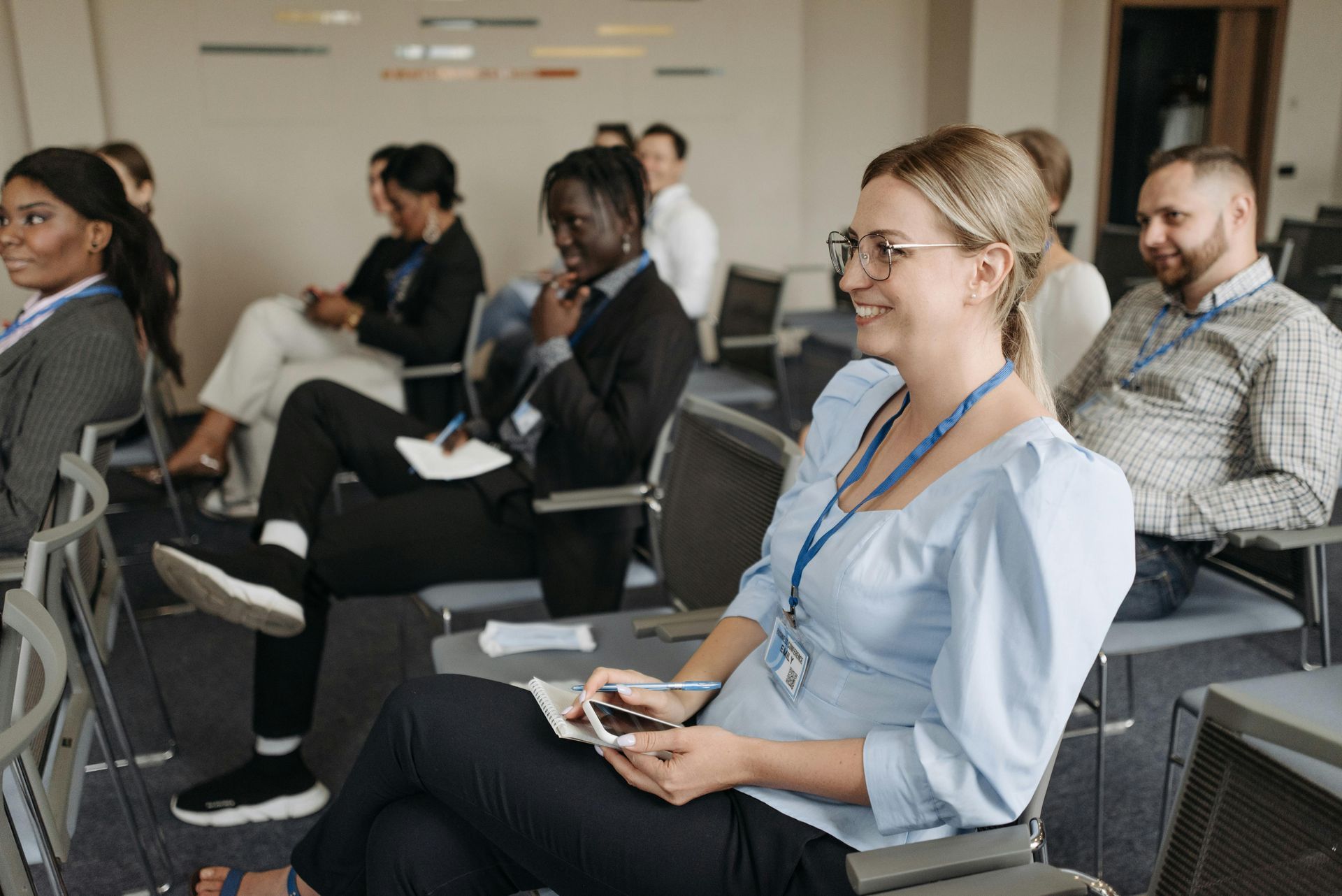 People attending a conference, seated and listening. A woman smiles, holding a phone. Others look on, attentive.