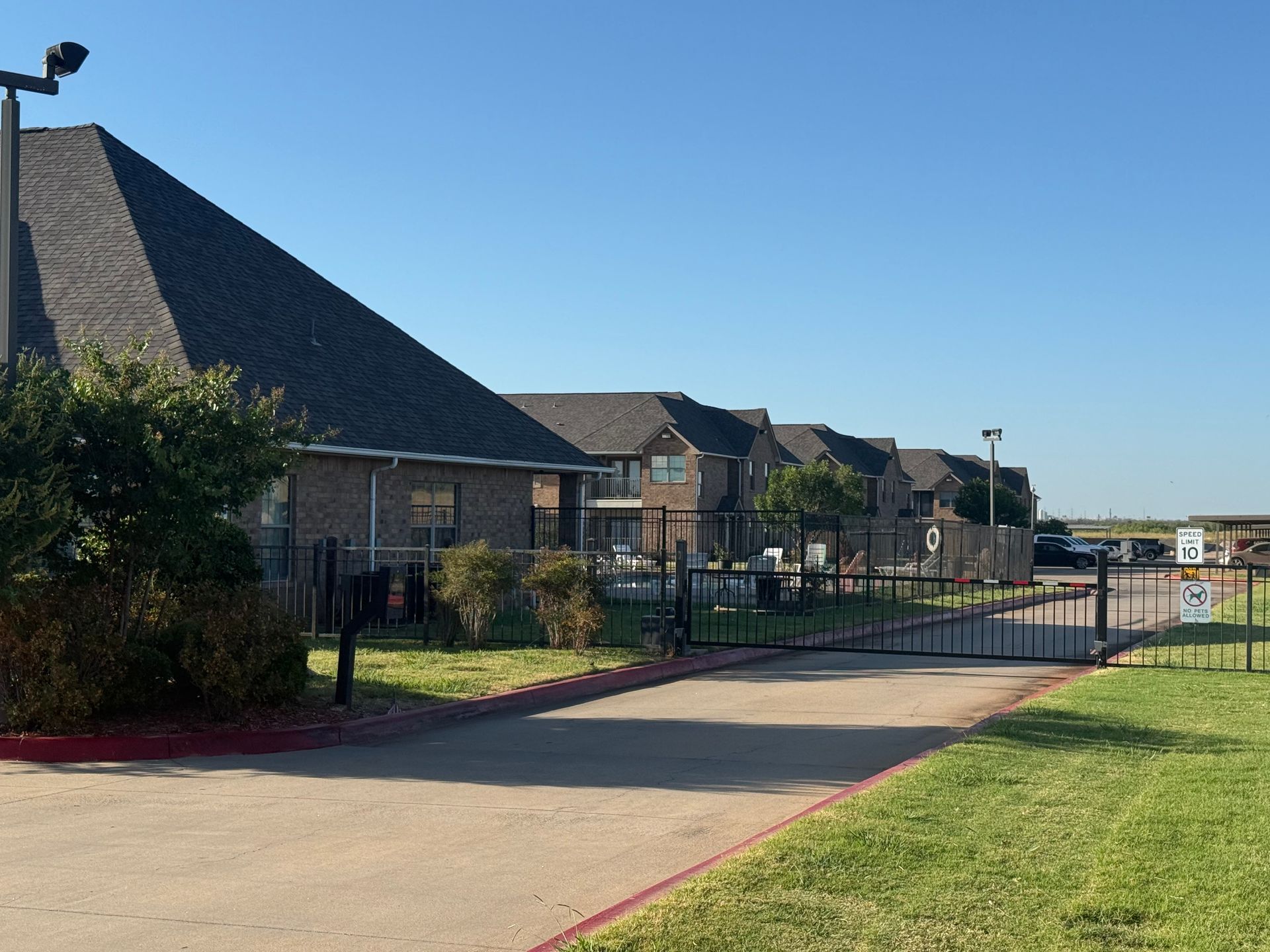 Apartment complex with a gated entrance on a sunny day. Brown brick buildings, brown roof, green grass and blue sky.