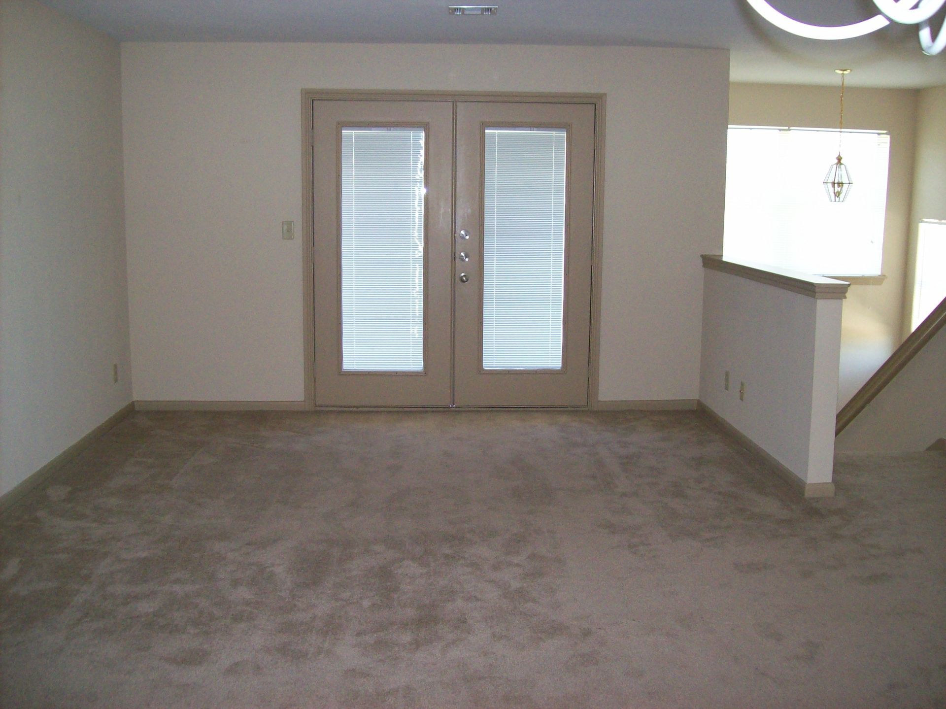 Empty room with beige walls, carpet, and French doors. Staircase on right.