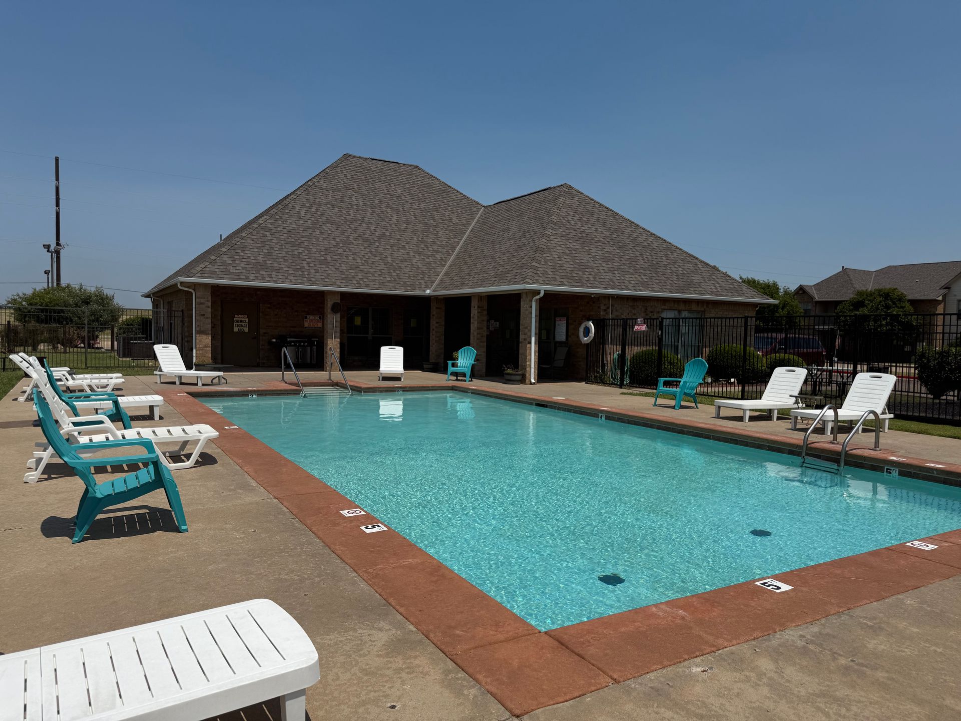 Pool with blue water and brick border, surrounded by lounge chairs, a building with a brown roof in the background.