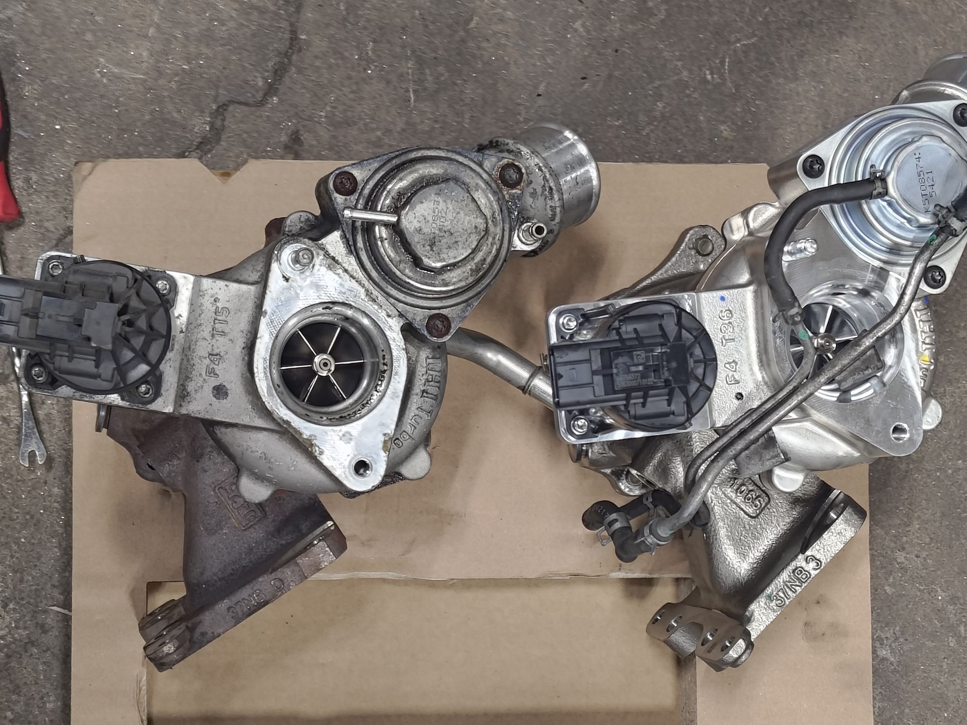 Two automotive turbochargers on a cardboard surface. Silver metal with various components, showing the internal turbine | La Saga Auto