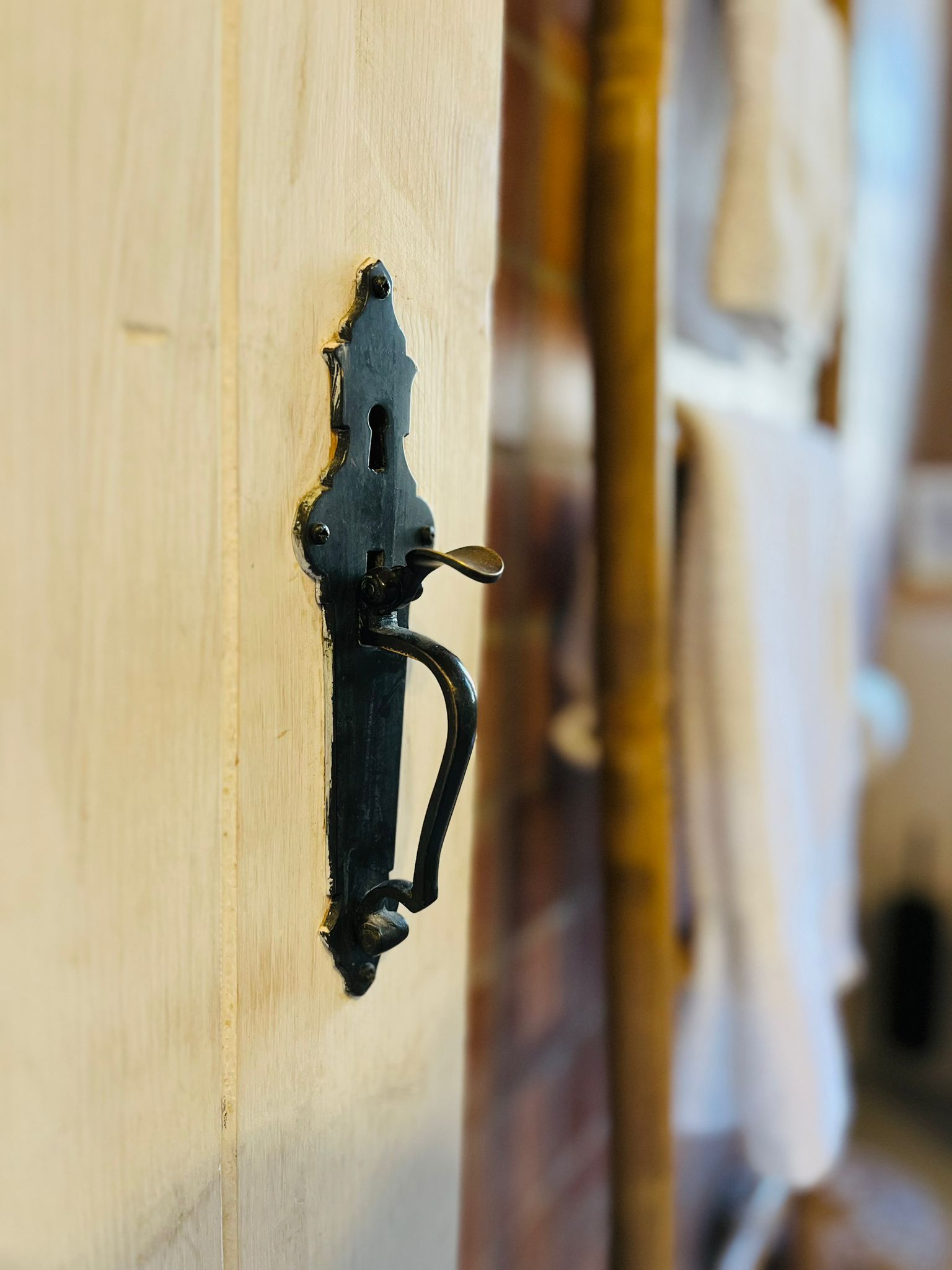 Black ornate door handle on a light wooden door. White towel and wooden post blurred in the background.
