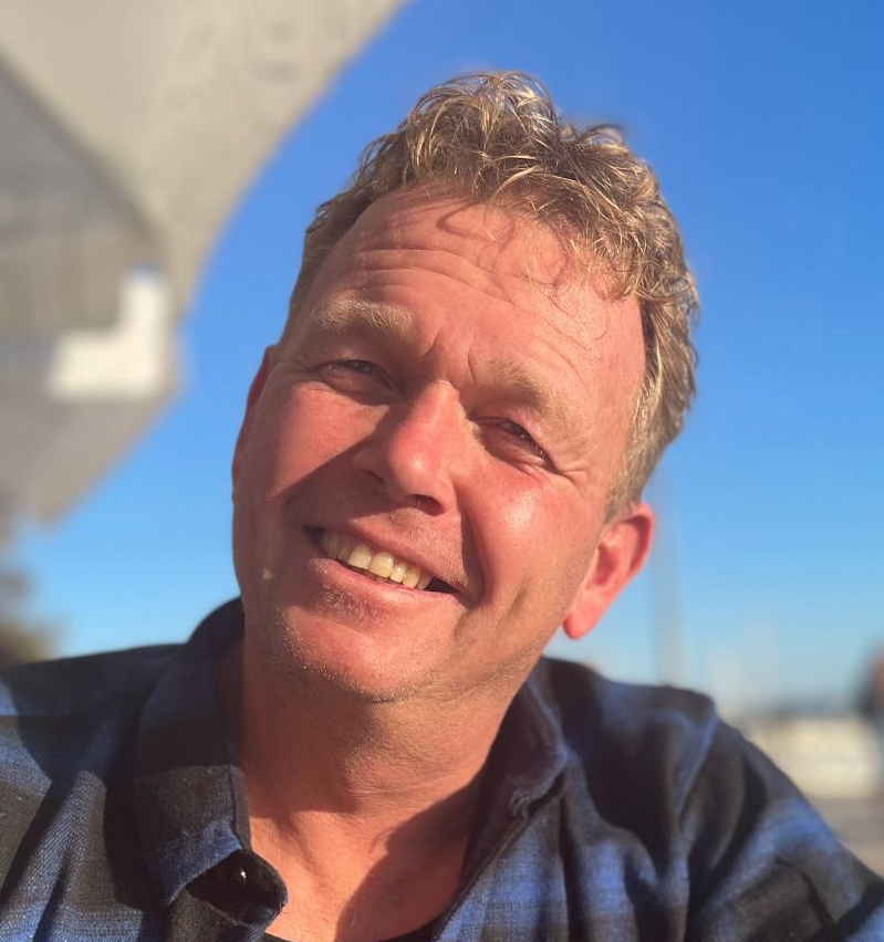 Smiling man with blond hair in blue plaid shirt, bright blue sky background.