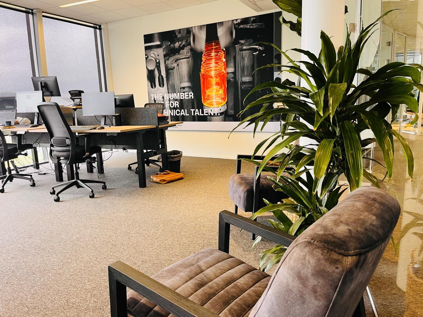 Office interior with desks, chairs, plants, and a large wall art with text.