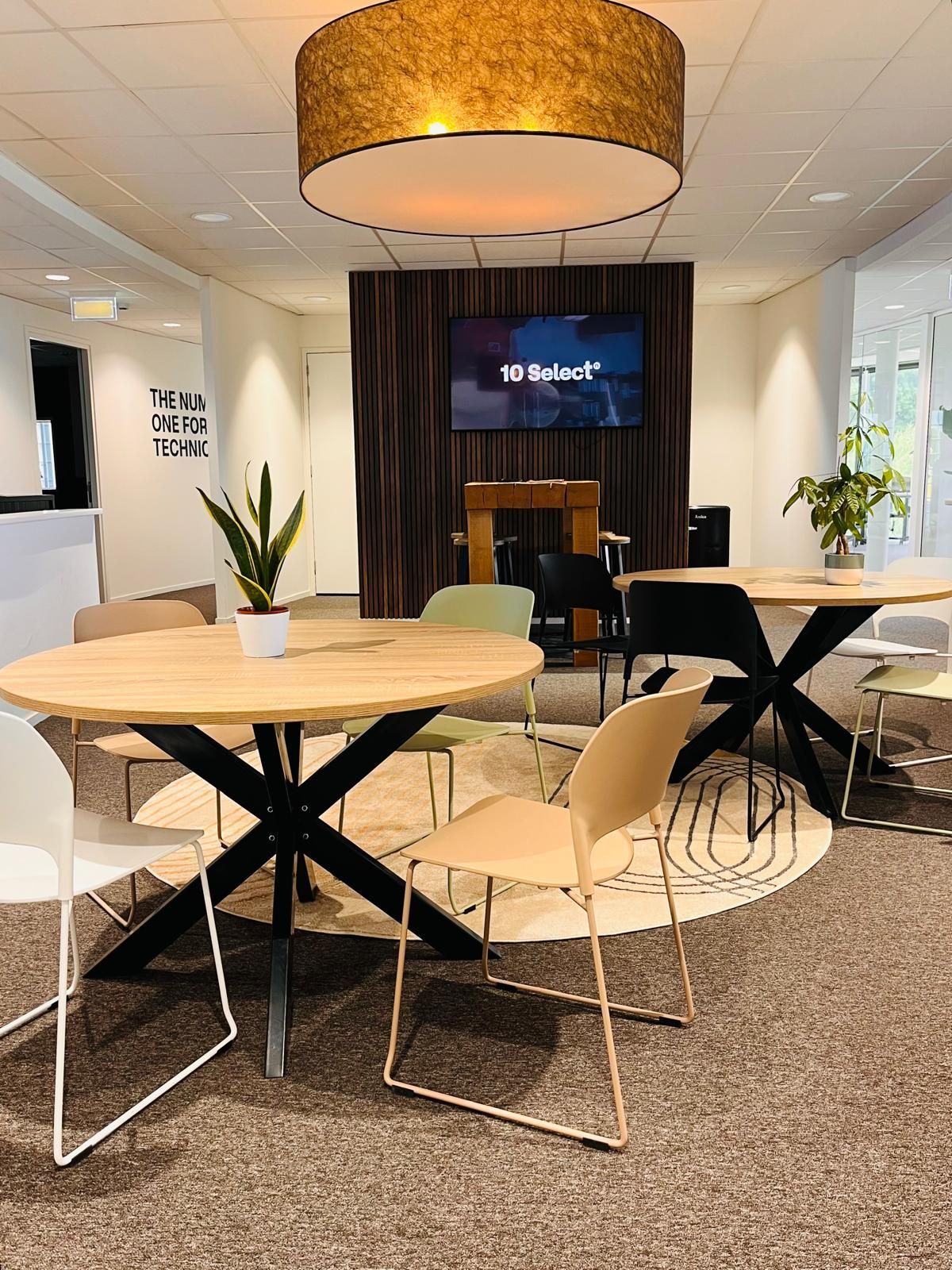 A modern office lounge with round tables, chairs, plants, and a TV. Warm lighting and neutral colors.