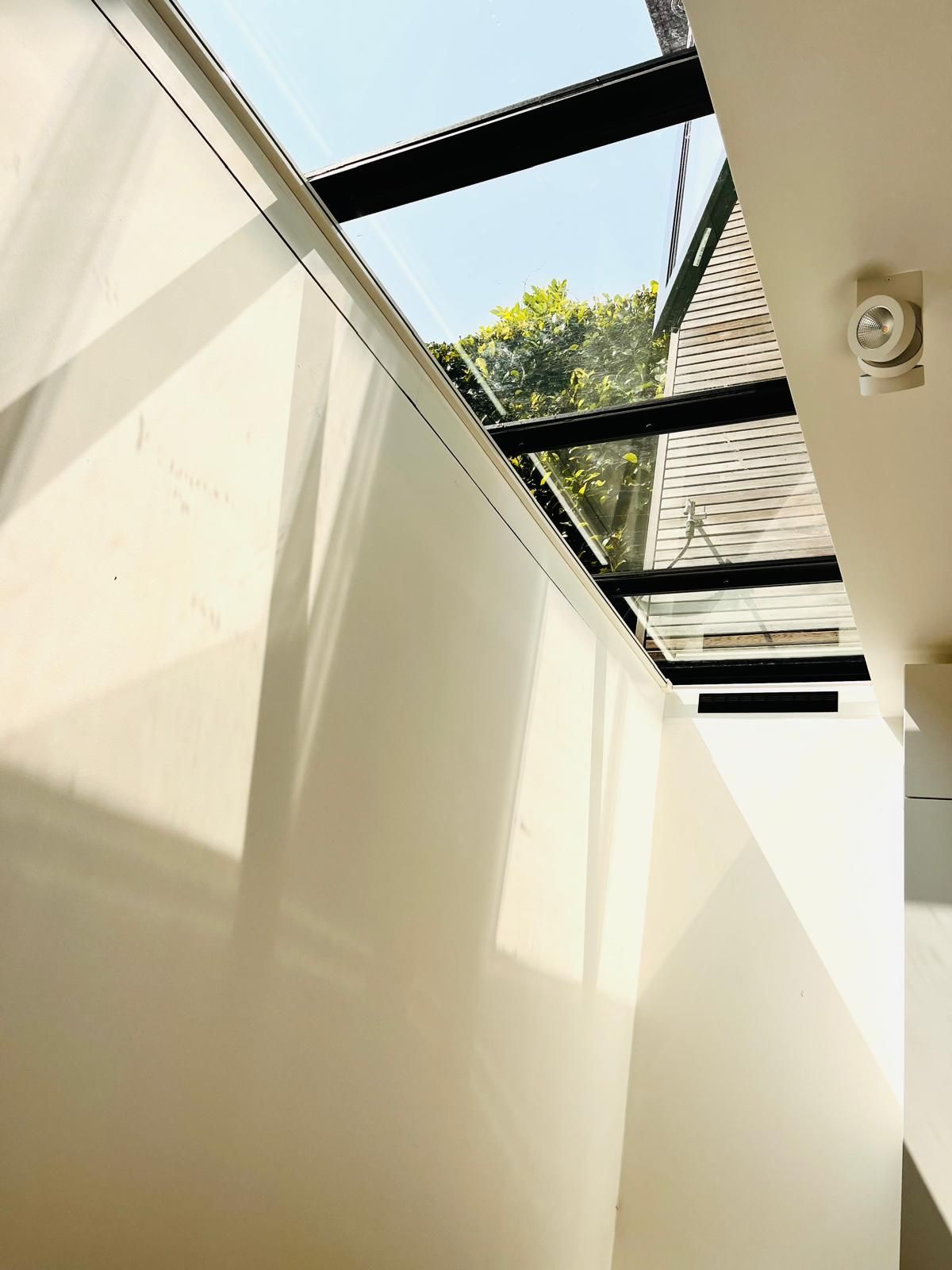 Sunlit interior with a glass roof, white walls, and a glimpse of trees outside.