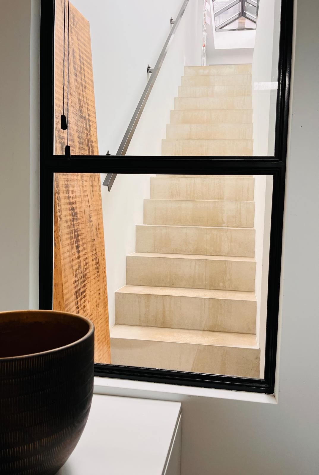 Staircase viewed through a black-framed window; light-colored steps ascend, wooden door to left, bowl in foreground.