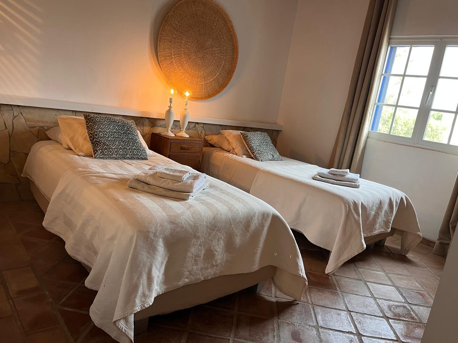 Two twin beds in a room with a window, lit by candles and a woven wall decoration.