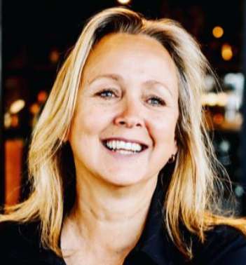 Smiling blonde woman, fair skin, dark top, natural light, blurred background.