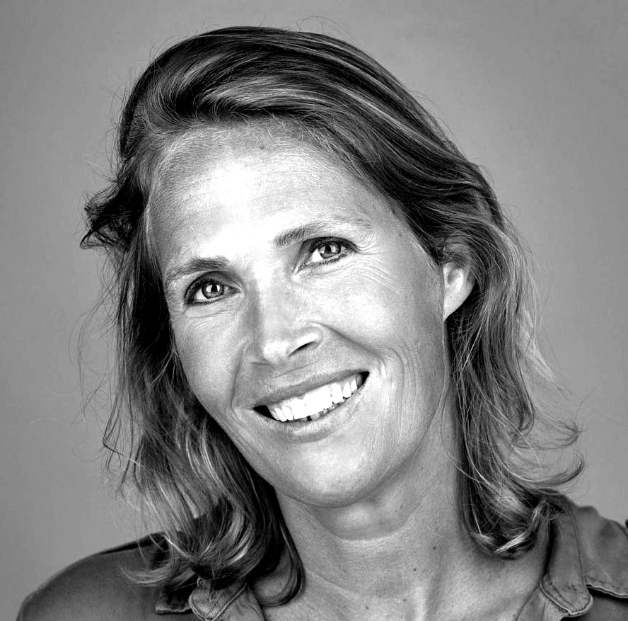 Woman smiling, looking slightly off-camera. Short, light brown hair, wearing a collared shirt, indoors.