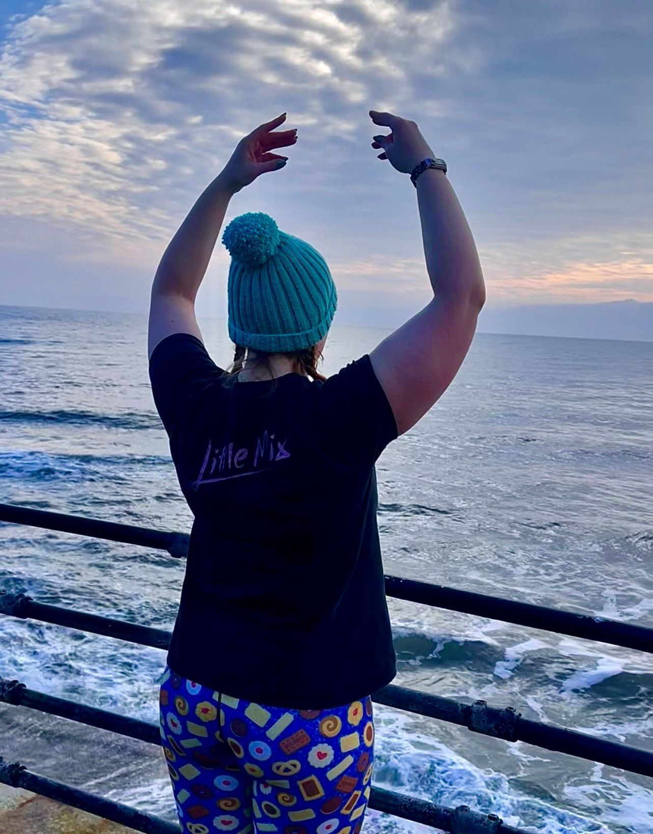 A woman wearing a black shirt and a blue hat stands on a pier overlooking the ocean