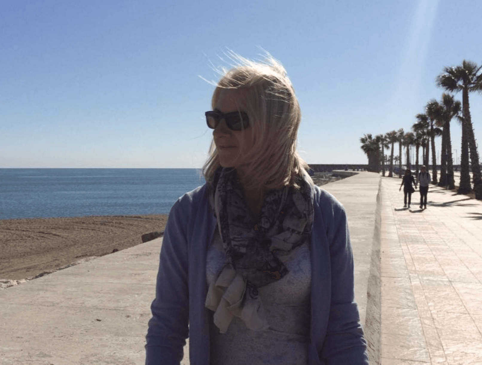 A woman wearing sunglasses stands on a sidewalk near the ocean