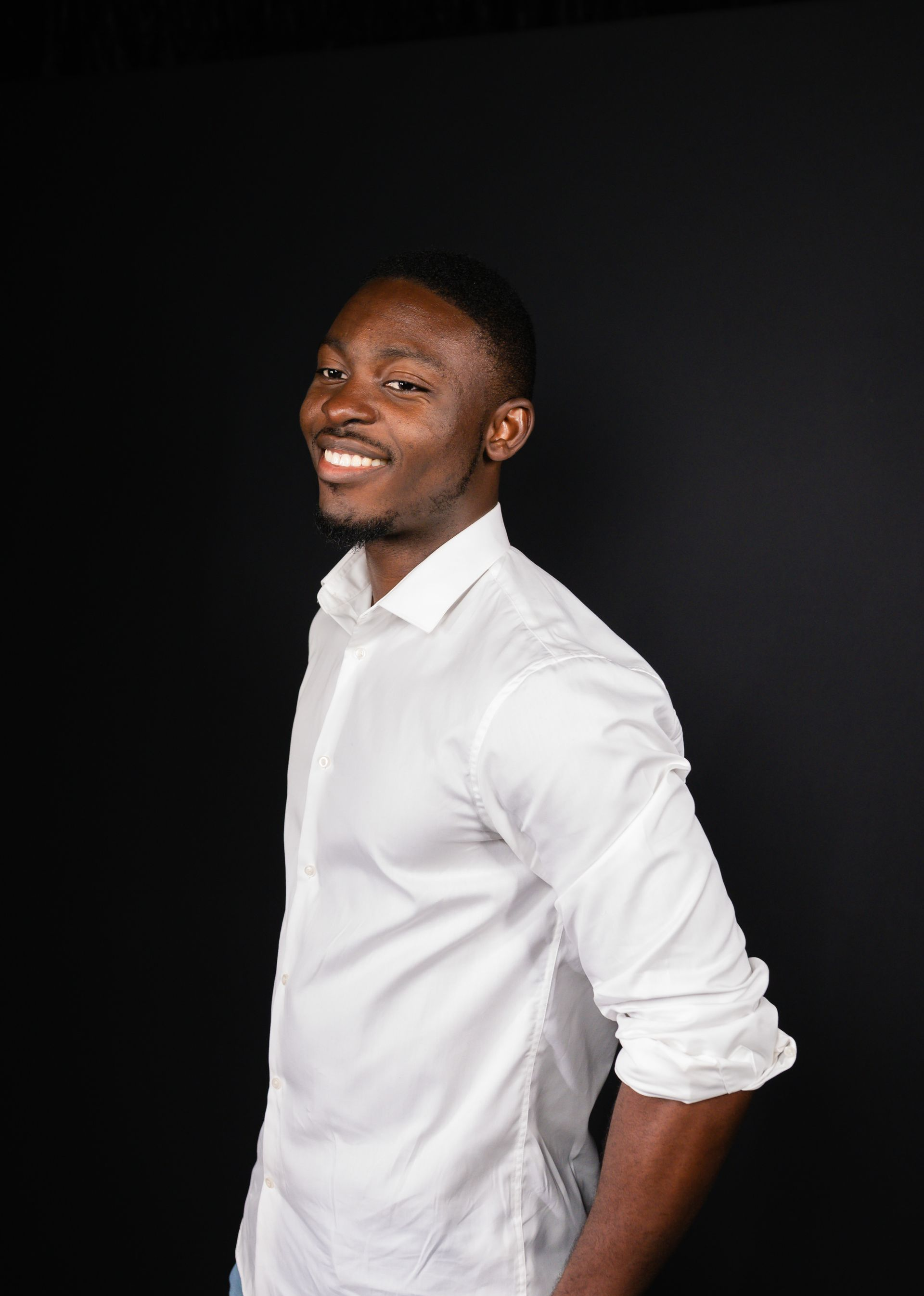 Man in white button-down shirt smiles, looking over his shoulder against a black background.
