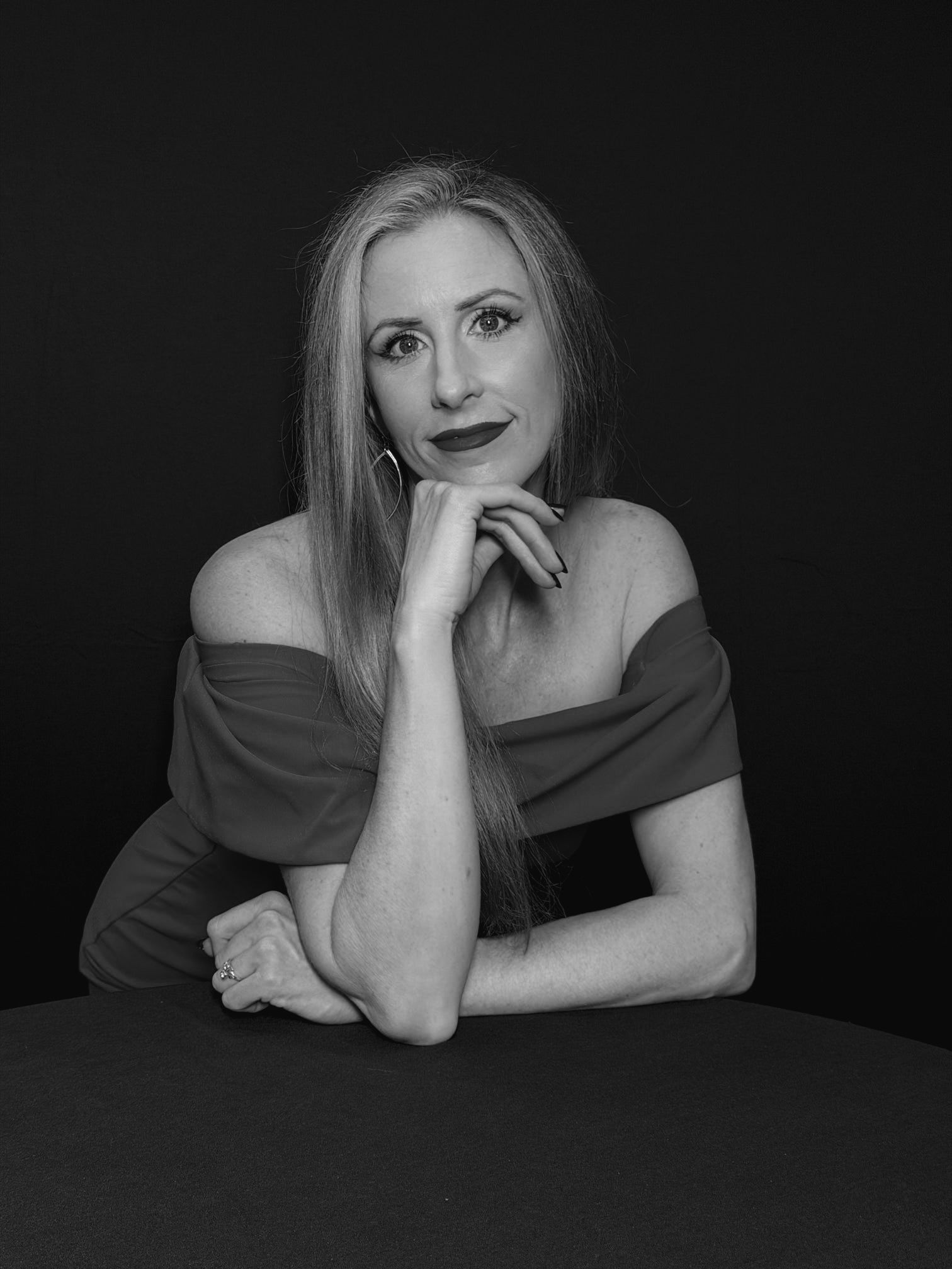 Woman in a red off-the-shoulder dress leans on a dark surface, hand to chin, against a black backdrop. Studio Headshot Photography