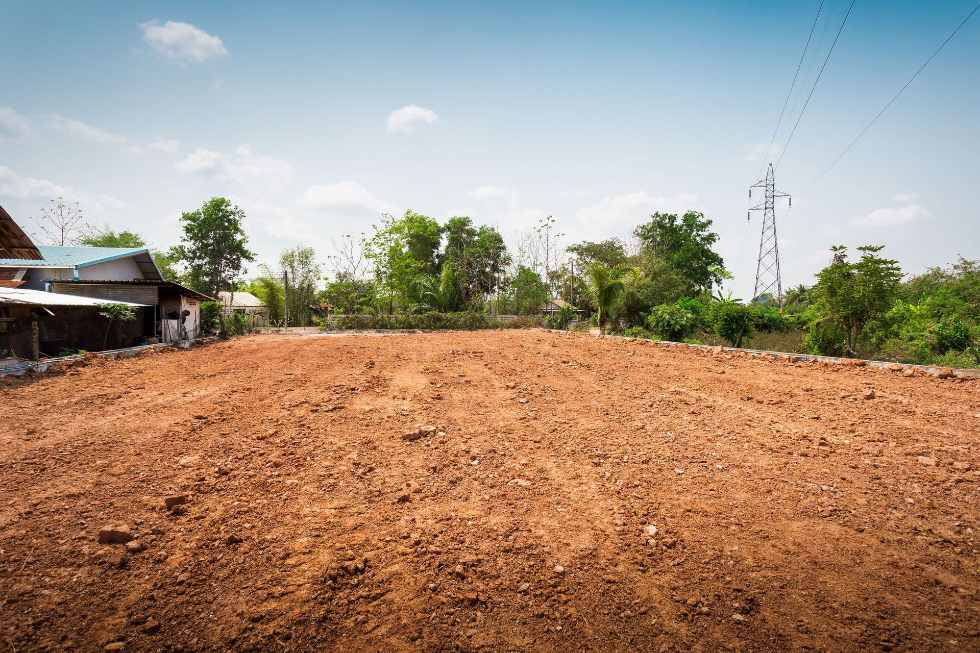 Cleared, reddish-brown earth plot, ready for construction, with trees and a house in background.