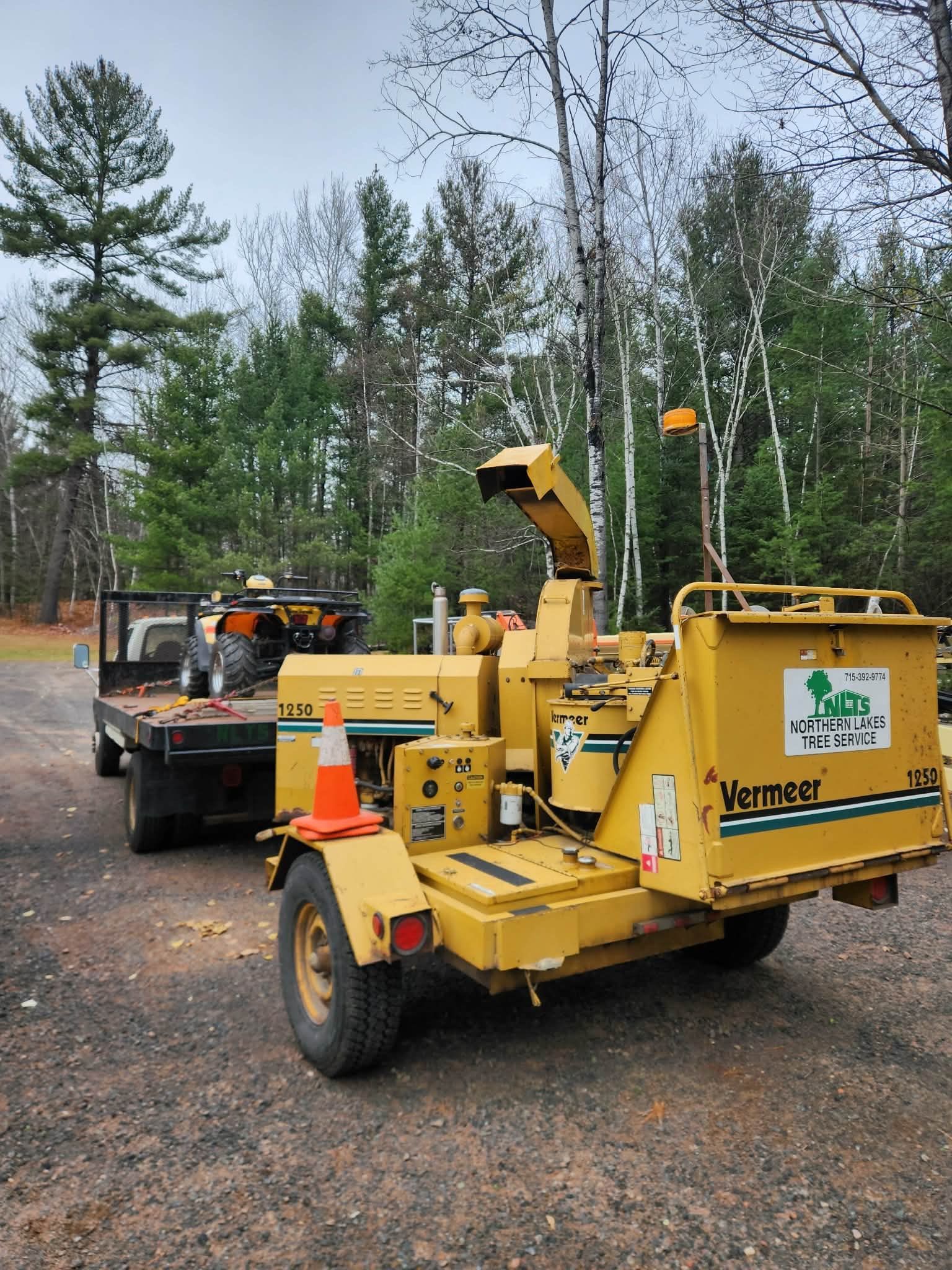 Person in a lift bucket trimming a tree branch in front of a house. Another person stands on the ground nearby.