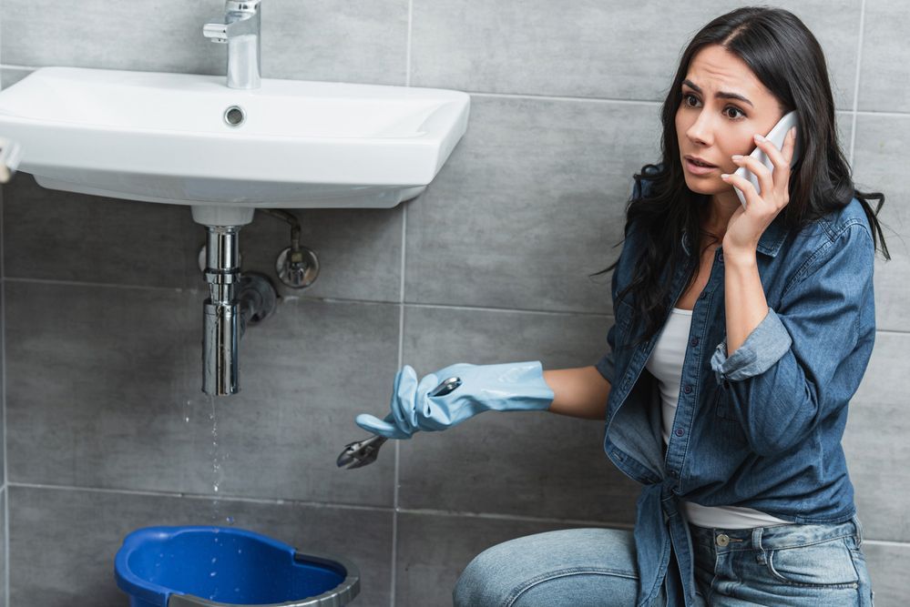 A woman is sitting under a sink talking on a cell phone.