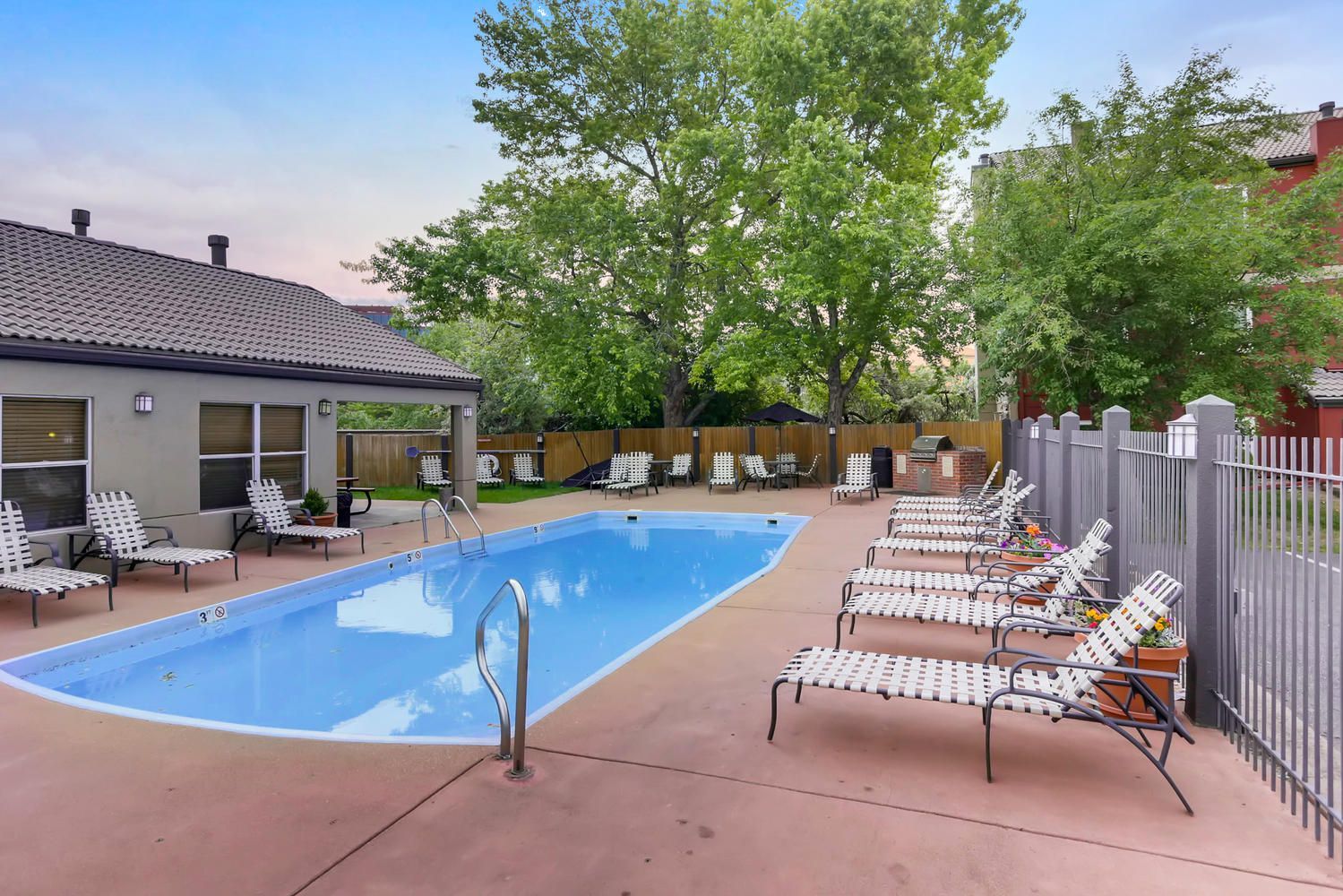 Pool with lounge chairs on a concrete patio, shaded by trees and a building.