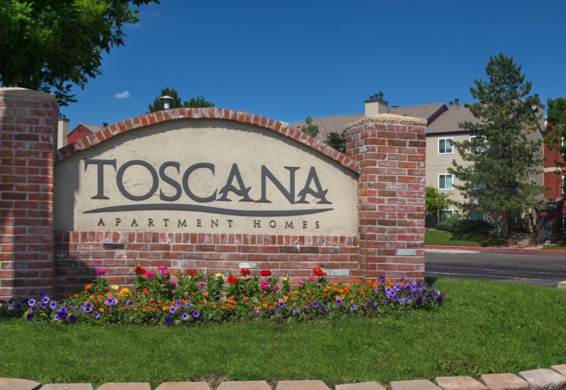 Sign reading 'Toscana Apartment Homes' at a brick entrance with flowers and landscaping.