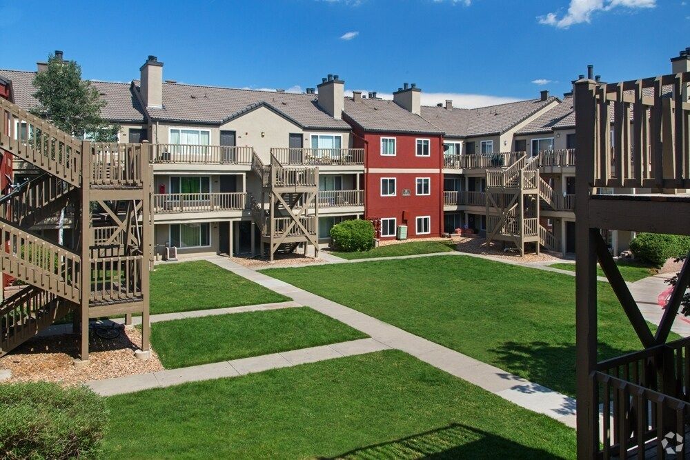 Apartment complex with a central green courtyard. Buildings have brown and red exteriors, with wooden stairs.