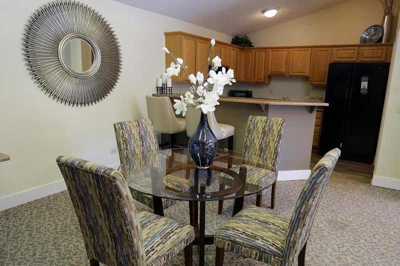 Dining area in an apartment: round glass table with six patterned chairs, vase centerpiece, and a kitchen in the background.