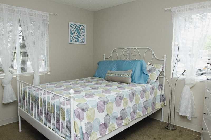 Bedroom in an apartment with a white metal bed, multicolor quilt, and sheer-curtained windows.