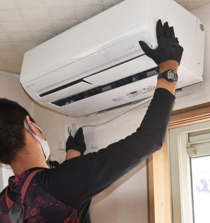 A technician wearing gloves and a face mask installs a white indoor air conditioning unit on a wall near a window.