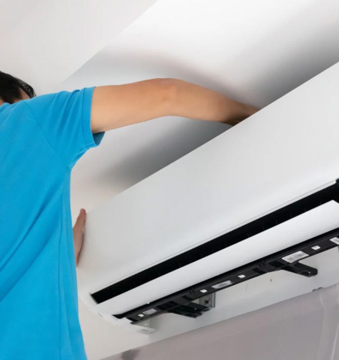 A person in a blue shirt reaches up to install or service a white wall-mounted air conditioner unit.