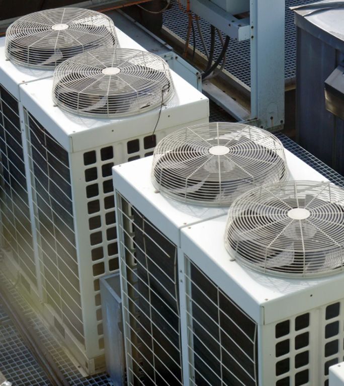 A high-angle view of four white, square outdoor HVAC condenser units with circular wire fan guards on a rooftop.