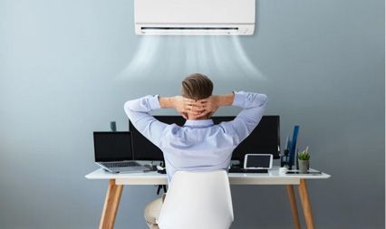 A person sitting at a desk with arms behind their head, relaxing in front of a wall-mounted air conditioner.