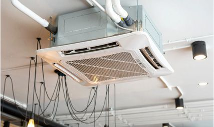 A white square ceiling-mounted cassette air conditioner unit installed in a room with industrial-style exposed lighting.
