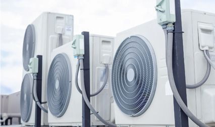 A row of four off-white outdoor air conditioning units with circular fans, mounted on a rooftop under a bright sky.