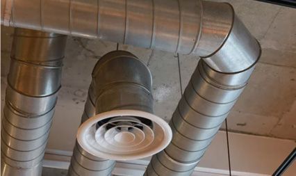 Metal ventilation ducts connected to a white ceiling-mounted circular air diffuser in an industrial-style interior.