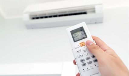 A hand holding a white air conditioner remote control aimed at a wall-mounted AC unit.