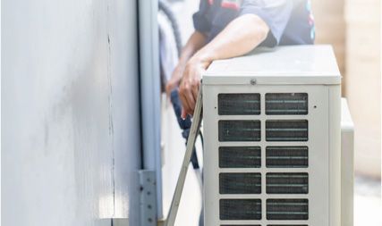 A technician in a blue uniform services the outdoor unit of an air conditioner next to a wall.