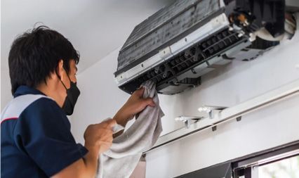 A worker wearing a face mask wipes the interior of a wall-mounted air conditioner unit with a cloth.