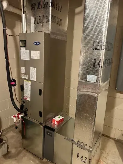 A Carrier furnace unit with silver metal ductwork installed against a beige concrete block wall in a basement.
