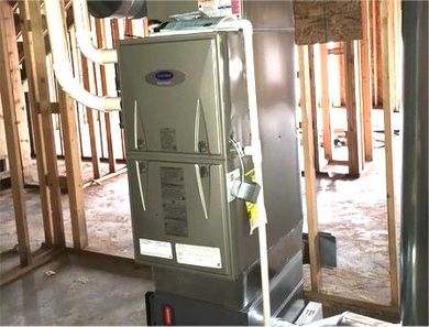 A grey Carrier furnace unit with white PVC piping, installed inside an unfinished room with wooden wall studs.