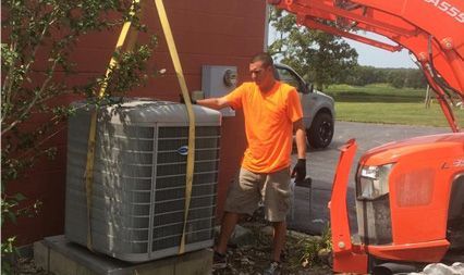 A worker in an orange shirt guides a large AC unit being lifted by a tractor near a building.