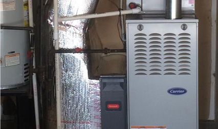 A silver Carrier furnace stands beside a water heater in a room with silver insulation panels on the walls.