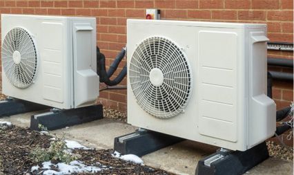 Two off-white air-source heat pump outdoor units sitting on black mounting blocks against a red brick wall.