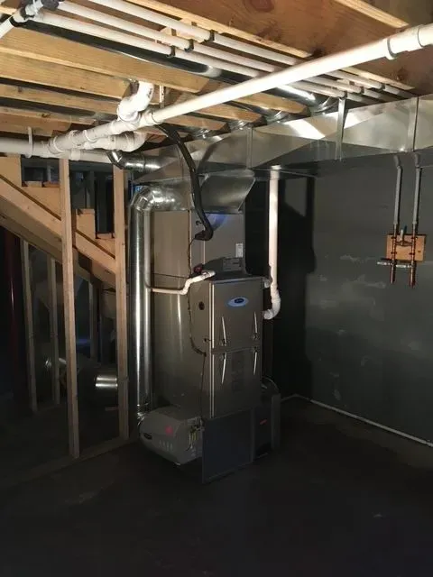 A silver gas furnace stands in a basement with exposed wooden ceiling joists and various white PVC and metal ductwork.