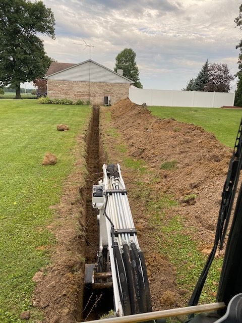View from inside an excavator cab showing a narrow trench being dug through a grassy yard toward a brick building.