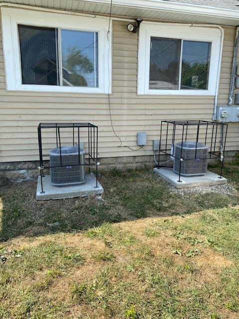 Two air conditioning units sit on concrete pads outside a beige house, each enclosed in a black metal protective cage.