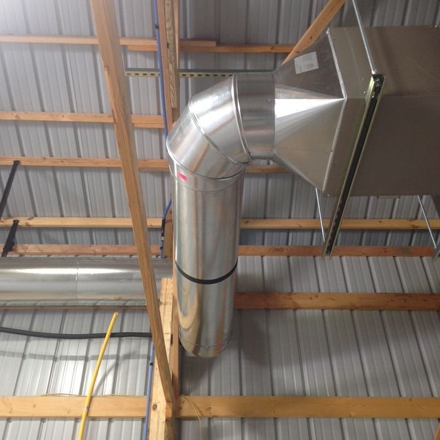 Metal ductwork transitions from a rectangular vent to a circular pipe, hanging from the wooden rafters of a metal ceiling.