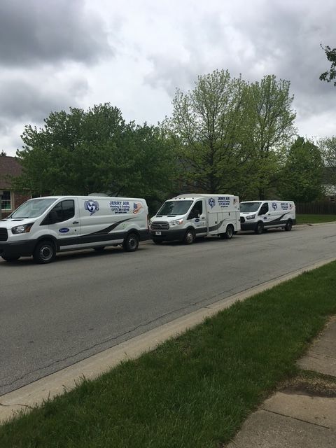 Three white work vans parked in a row along a suburban street lined with green trees on a cloudy day.