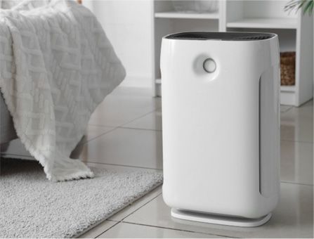 A white air purifier stands on a tiled floor next to a bed with a white blanket and a grey rug.
