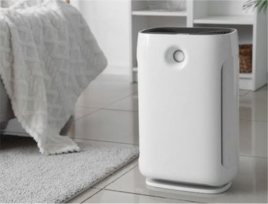 A white air purifier stands on a tiled floor next to a bed with a white blanket and a grey rug.